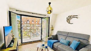 A living room with a grey sofa, a white furry rug, a glass coffee table, a TV on a wooden stand, and a view of buildings through a glass door. - Standing, Centre, 2 Sdb 2 WC, Balcon, Parking (Deauville)