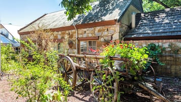 A rustic stone building with a metal roof, surrounded by greenery and a vintage wooden cart.