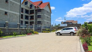 A multi-story building with a red roof, a paved driveway, and a parked car. - Isomer Comfort Homes (Nakuru)
