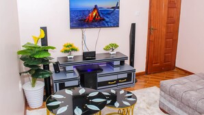 A living room with a flat-screen TV mounted on the wall, a black entertainment unit, a round coffee table with leaf patterns, a potted plant, and a cushioned armchair. - Isomer Comfort Homes (Nakuru)