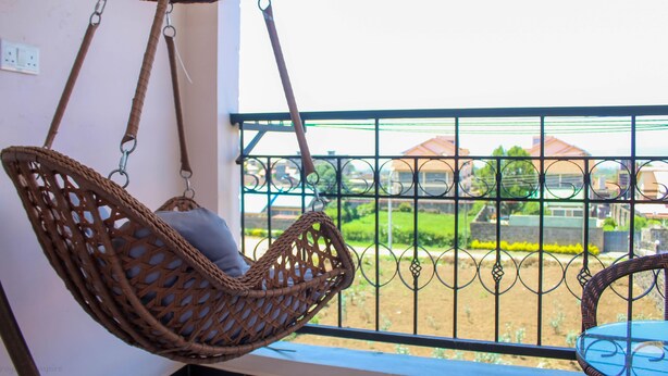 A wicker hanging chair with a cushion on a balcony with a metal railing, overlooking a residential area. - Isomer Comfort Homes (Nakuru)