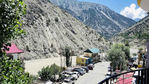 A mountainous landscape with a road leading to a small town. There are parked cars, buildings, and a few trees visible. - The Kuber Hotel & Restaurant (Puh)