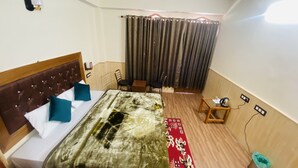 A hotel room with a bed, a small table, a chair, a window with curtains, and a wall-mounted light fixture. - The Kuber Hotel & Restaurant (Puh)
