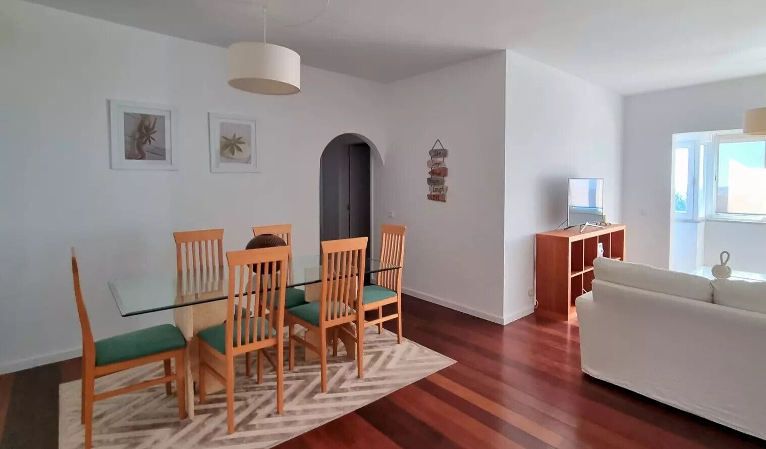 Uma sala de estar moderna com uma mesa de jantar de vidro, cadeiras de madeira, um sofá branco e um móvel para TV.