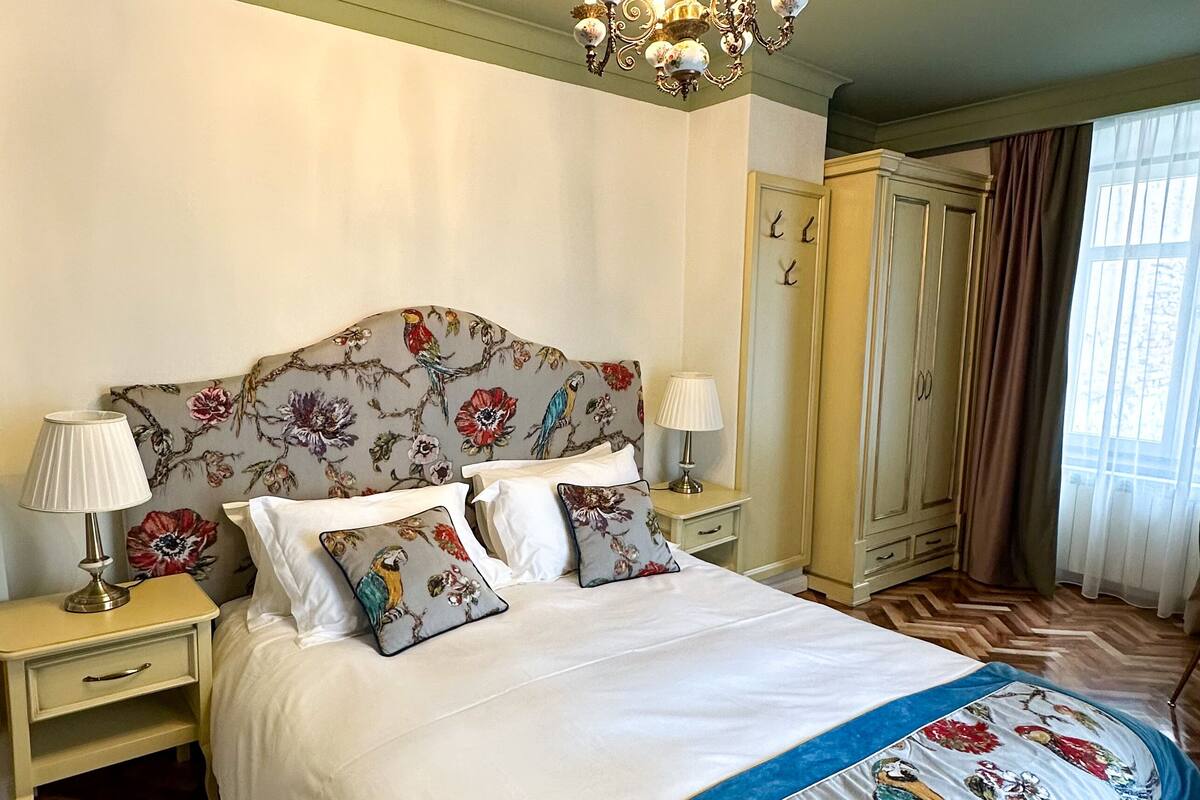A bedroom with a large bed, floral headboard, bedside lamps, a wardrobe, and a chandelier.