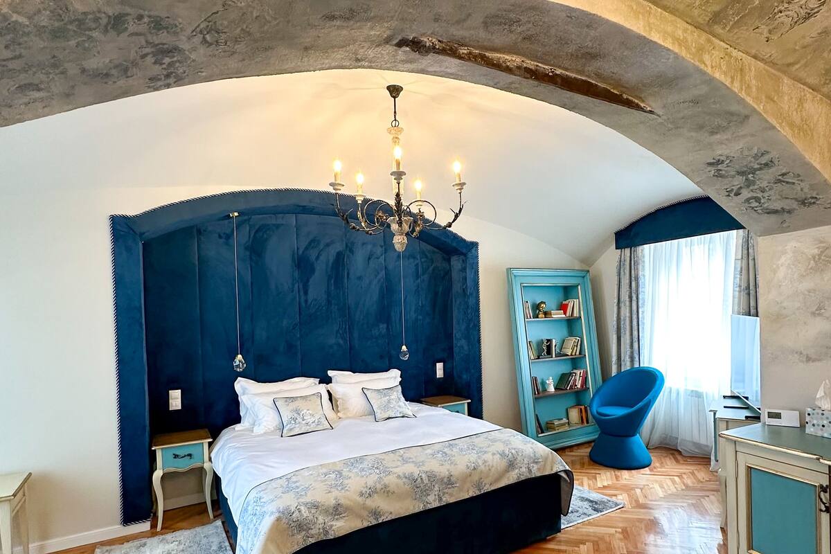 A bedroom with a large bed, a chandelier, a blue chair, and a bookshelf.