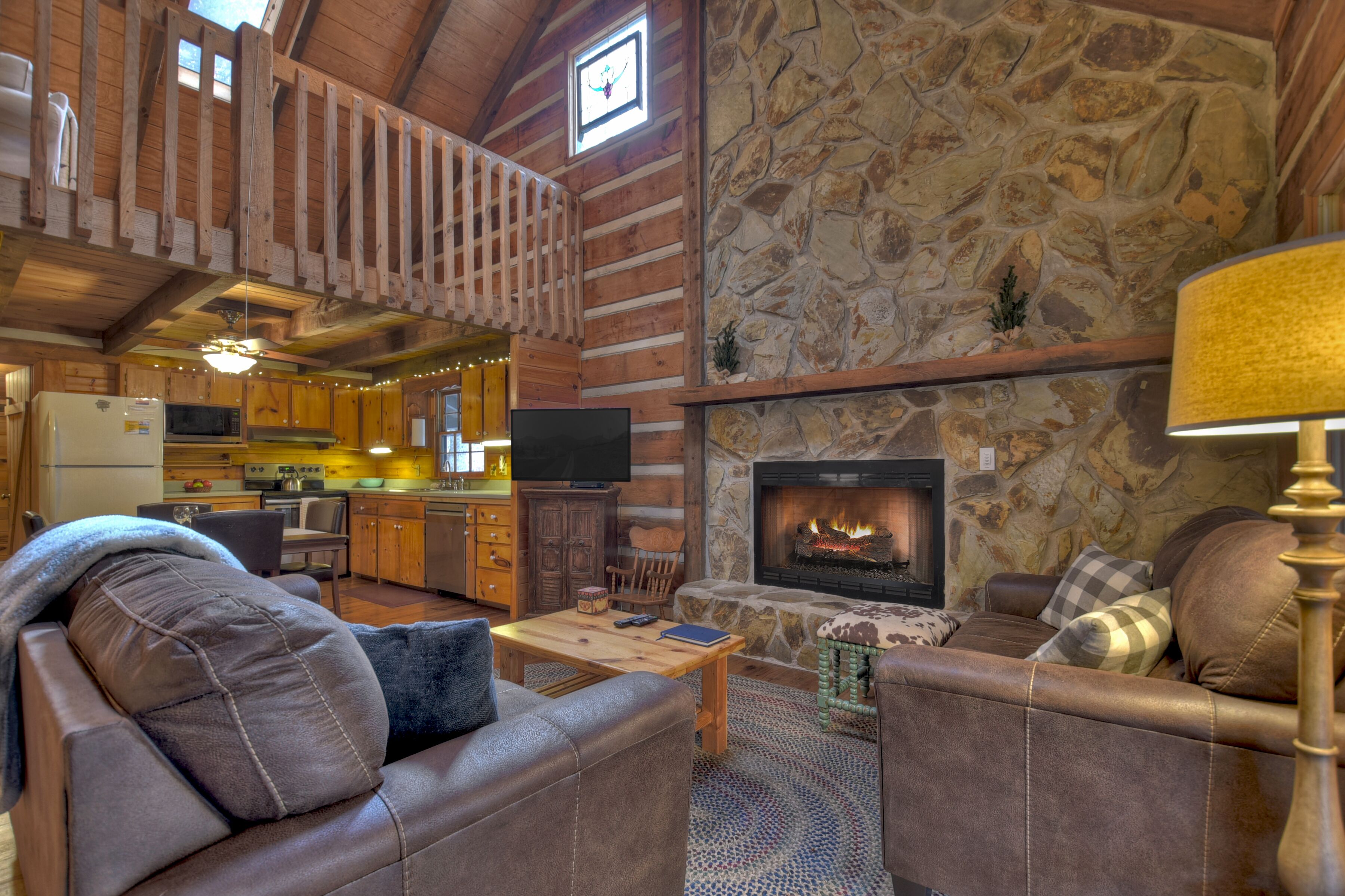 A log cabin interior with a stone fireplace, wooden furniture, and a kitchen area.