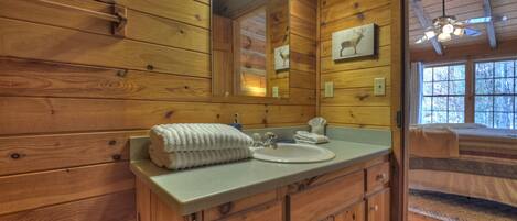 Une salle de bain rustique, aménagée dans une cabane en bois, avec un lavabo, un miroir et un lit donnant sur une fenêtre.