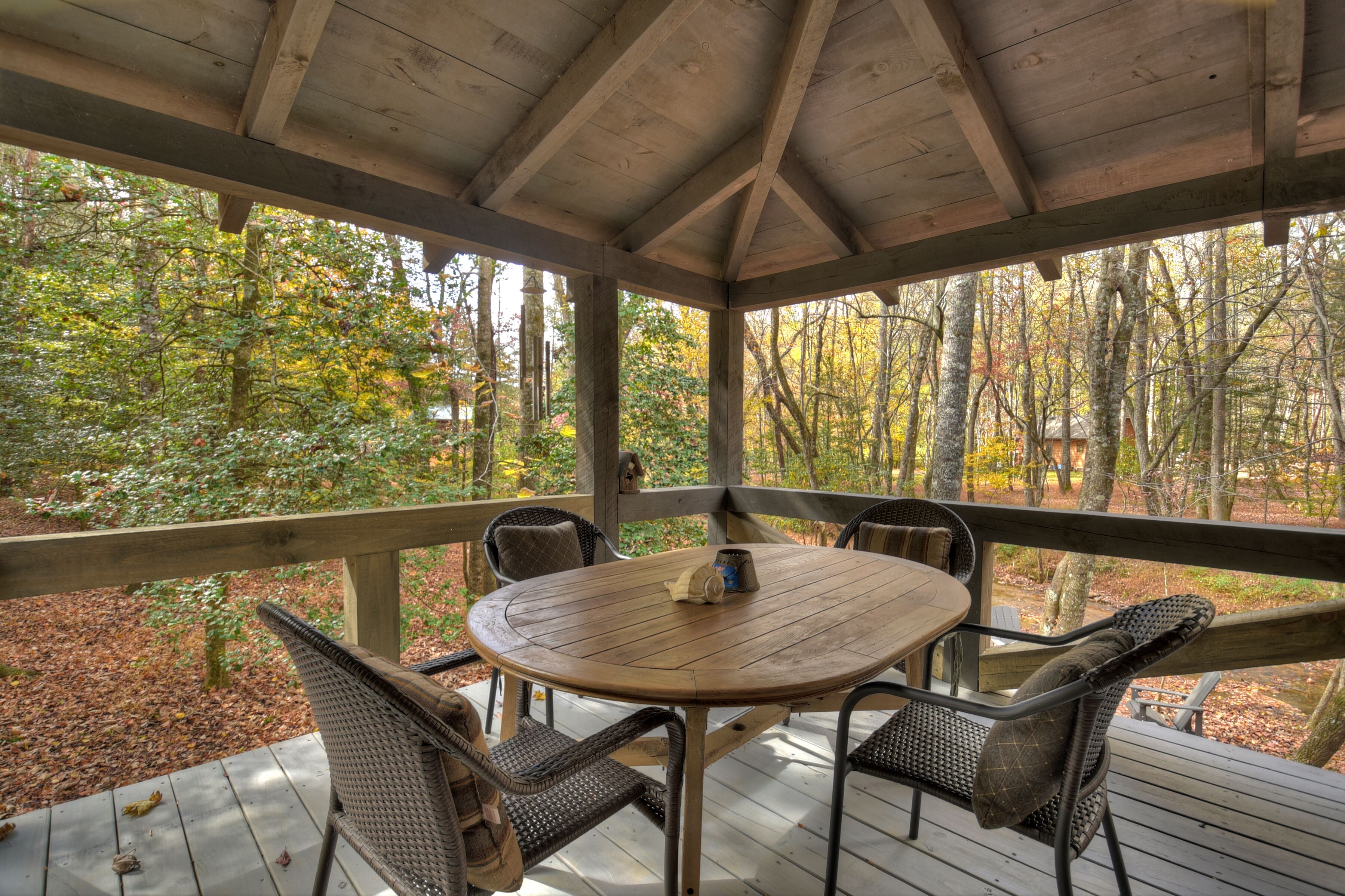 A covered outdoor seating area with a round table and wicker chairs, surrounded by a wooden deck and a forest backdrop.
