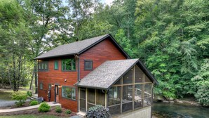 A two-story wooden house with a glass conservatory, surrounded by a lush green forest and a stream.