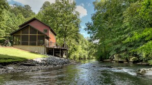A cabin with a deck overlooking a river, surrounded by dense forest.