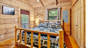 Ein Schlafzimmer einer Holzhütte mit Bett, Deckenventilator, Fenster mit Vorhängen und einem Wand-TV.