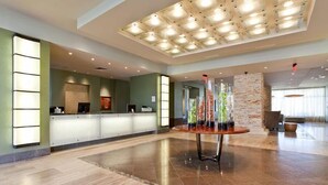 Un hall d’hôtel moderne avec un comptoir d’accueil, une table ronde agrémentée d’objets décoratifs et un plafond haut doté d’un éclairage encastré.
