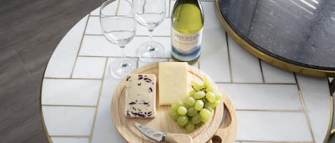 Una mesa redonda con superficie de azulejos blancos y dorados, una botella de vino, dos copas, una tabla de madera con queso y uvas, y un cuchillo.