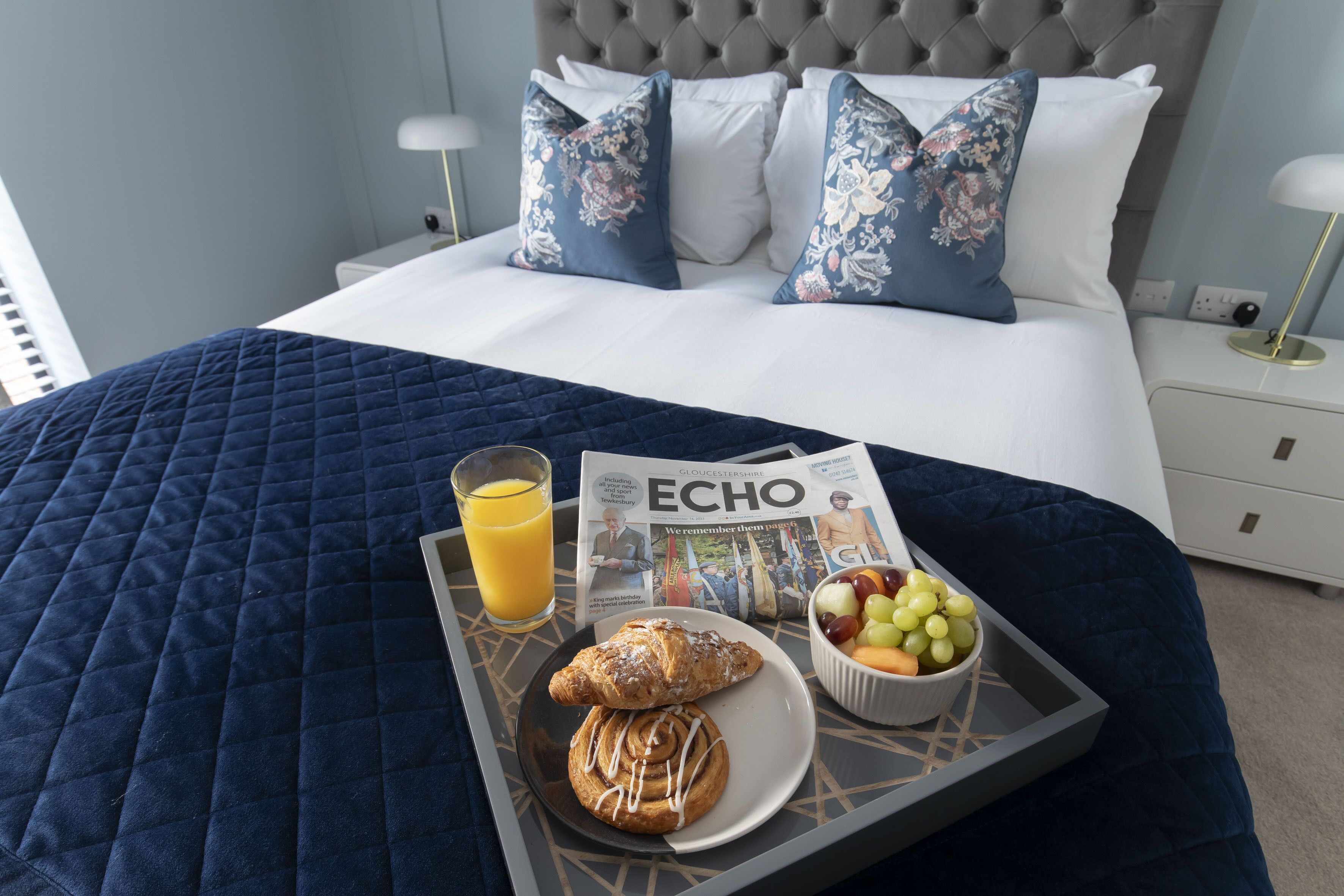 A neatly made bed with a blue quilt, a tray with croissants, a bowl of fruit, and a glass of orange juice.