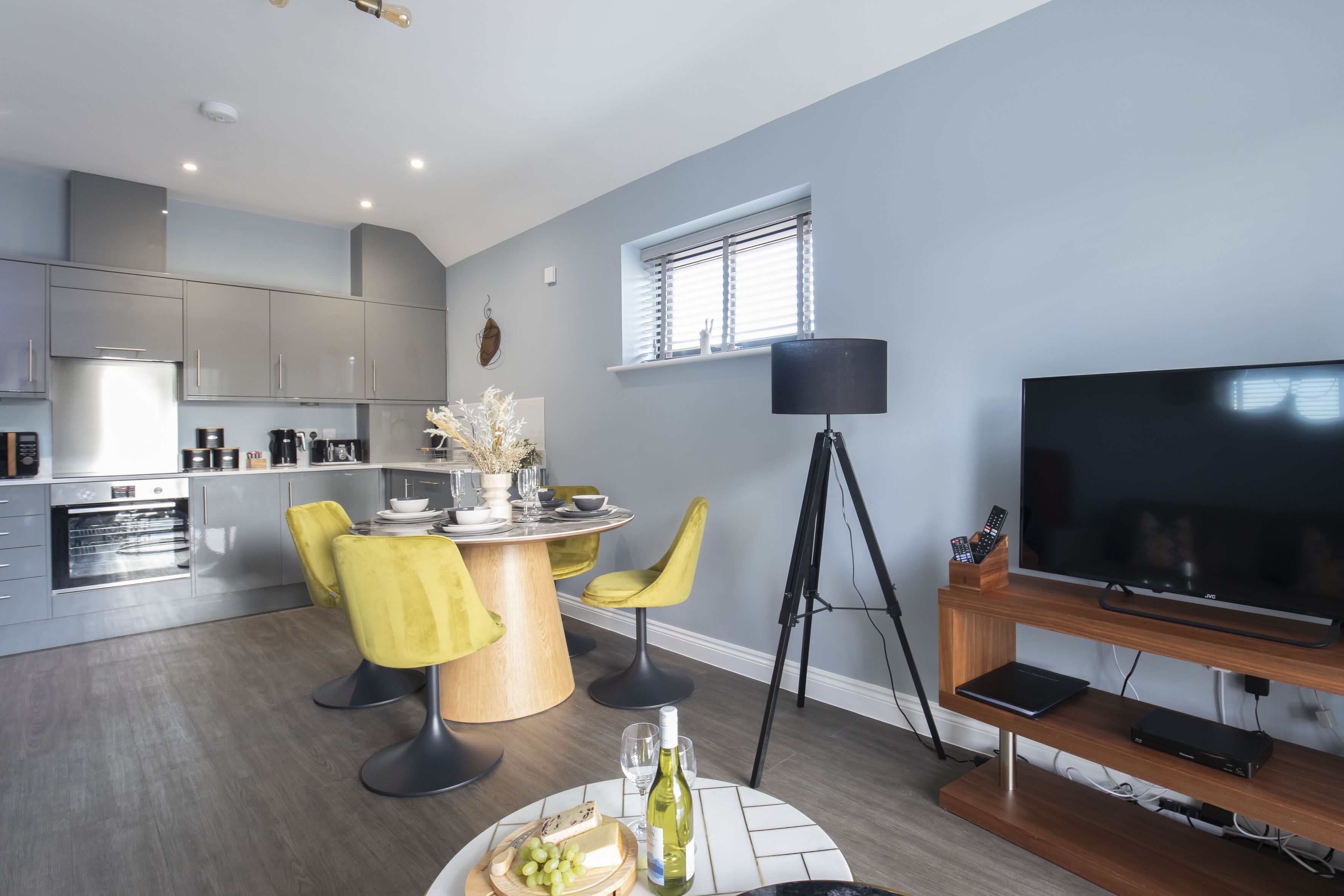 A modern kitchen with grey cabinets, a dining area with yellow chairs, a round table, and a TV on a wooden stand.