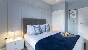 A bedroom with a tufted headboard, a bed with white bedding and blue floral pillows, a bedside table with a lamp, and a tray with breakfast items. - Elliot Oliver - Luxurious Two Bedroom Apartment in The Docks (Gloucester)