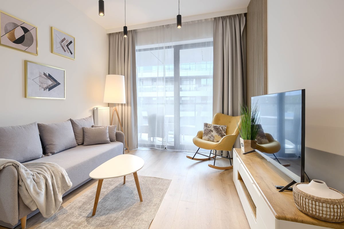 A modern living room with a grey sofa, a small white table, a rocking chair, a TV, and a large window with curtains.