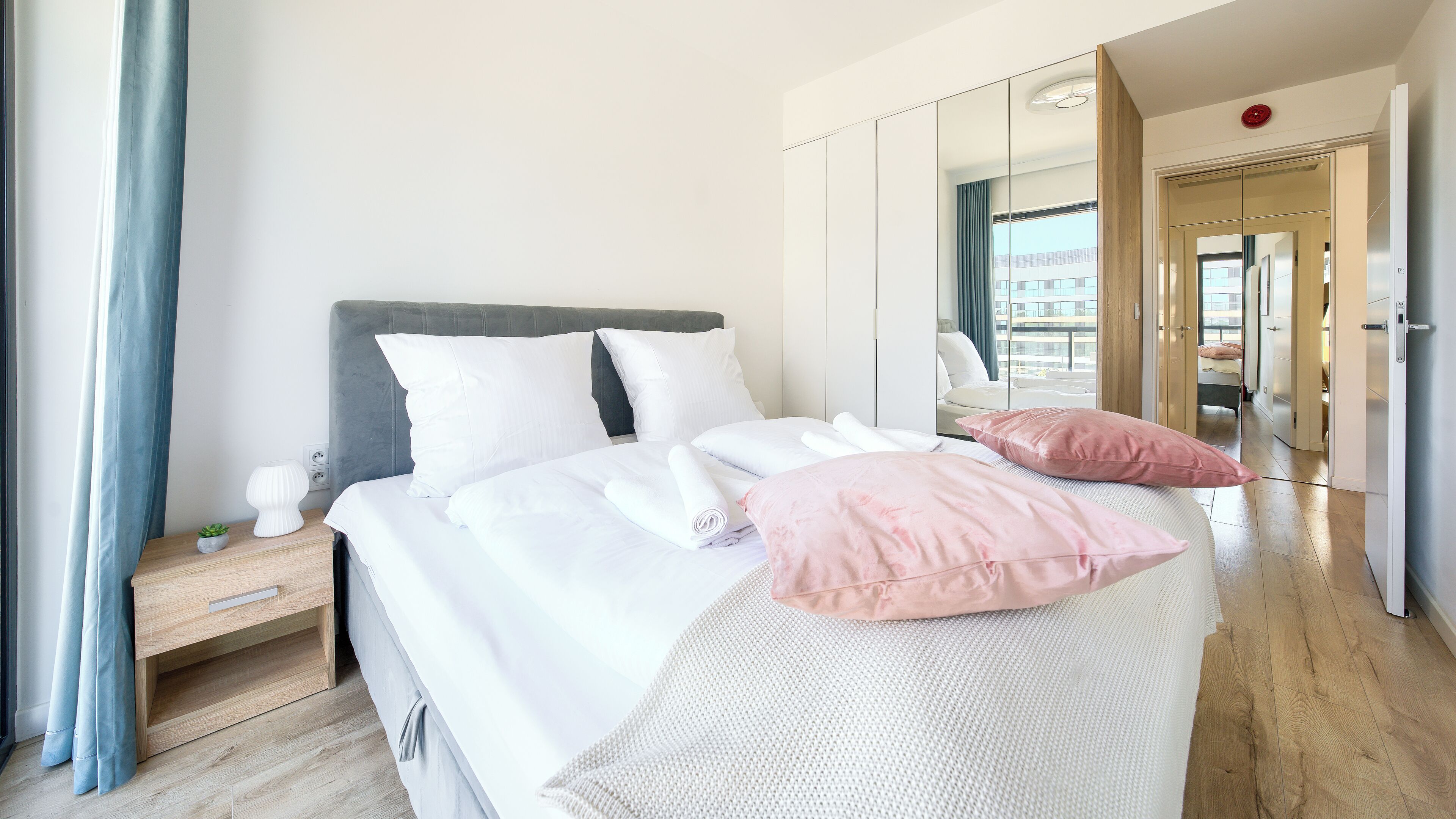 A neatly made bed with pillows, a bedside table with a lamp, and a large mirror on the wall.
