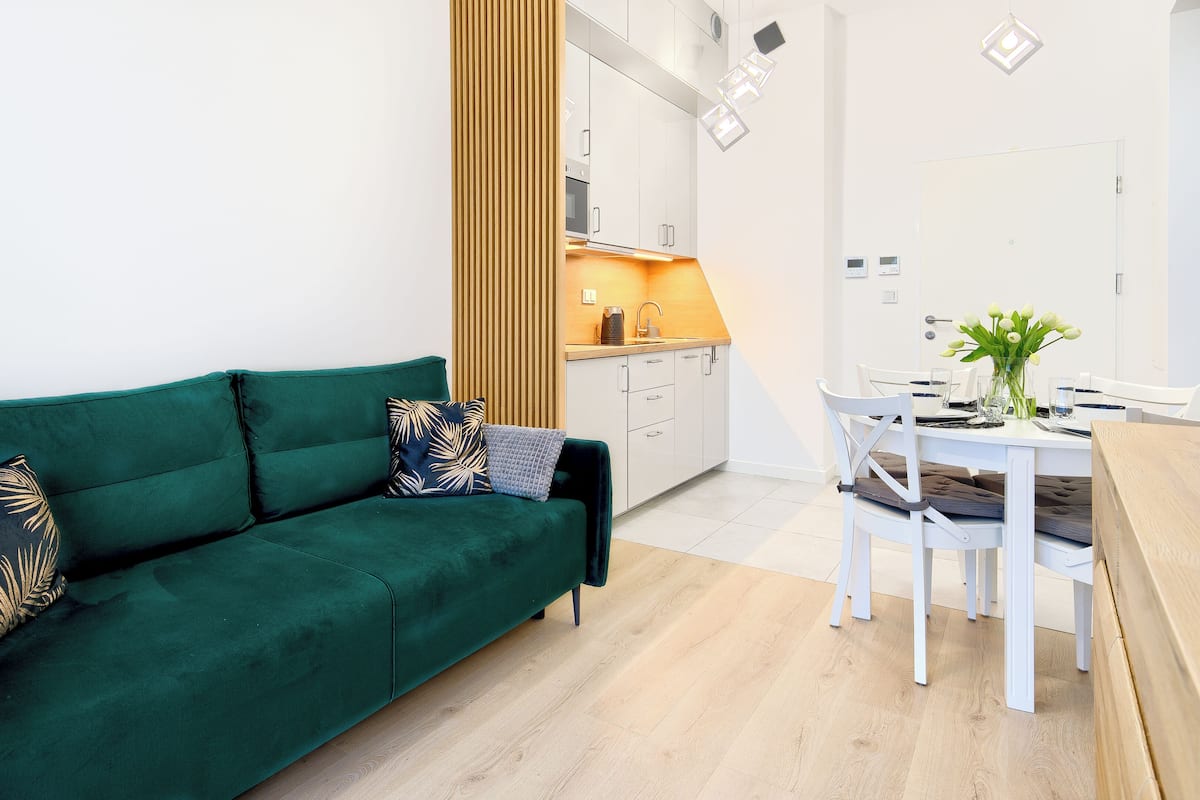 A modern living room with a green sofa, a dining table with chairs, and a kitchen area in the background.