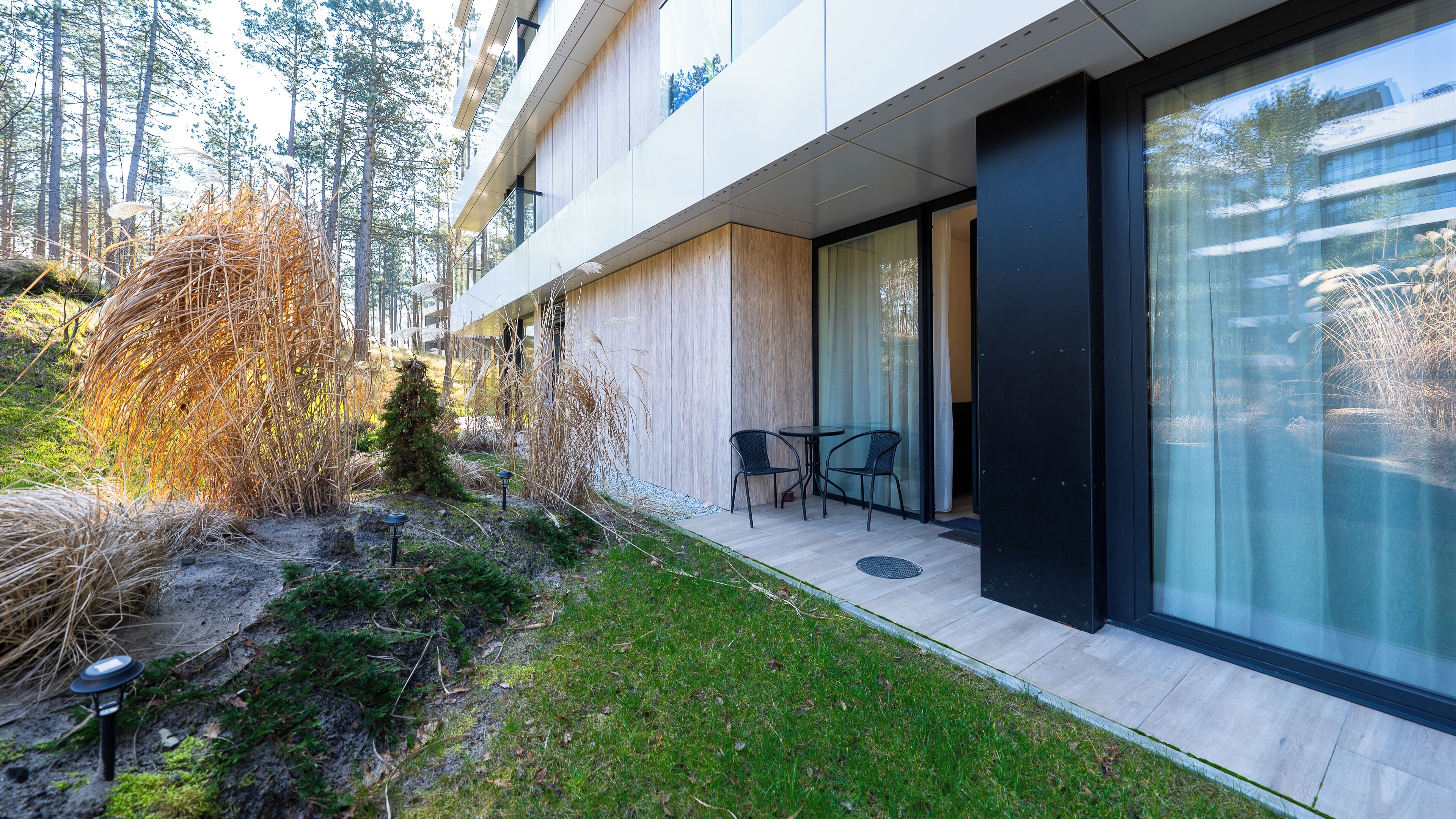 A modern building with a glass facade, a small outdoor seating area with chairs, and a well-maintained garden with grass and shrubs.