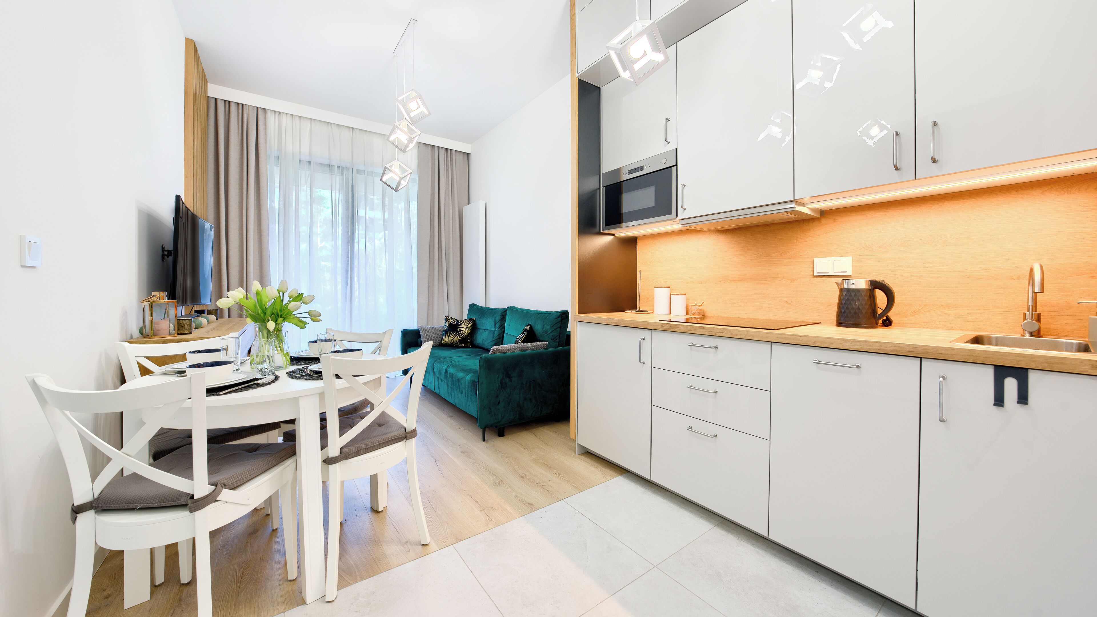 A modern kitchen with white cabinets, a microwave, and a sink. There is a dining area with a table and chairs, and a green sofa in the background.