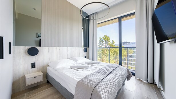 A modern bedroom with a large bed, a bedside table, a wall-mounted TV, and a view of the city through large windows. - Apartamenty Sun & Snow Shellter (Trzebiatow)