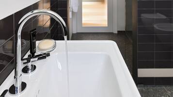 A modern bathroom with a white sink, black tiles, a round mirror, and a door leading to another room.
