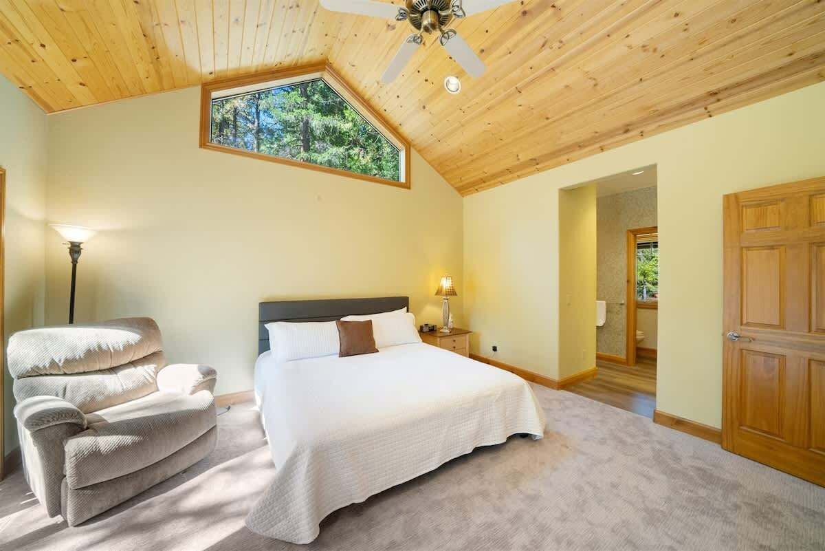 A bedroom with a large bed, a recliner, a ceiling fan, and a window with a view of trees.