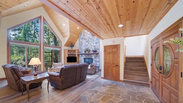 A spacious living area with a stone fireplace, wooden flooring, and large windows offering a view of the outdoors. - Wishram Retreat - 24 Wishram Trail (Graeagle)