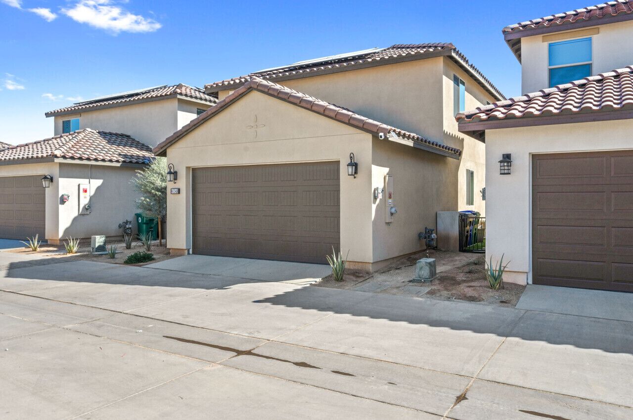 A row of residential houses with beige stucco walls and brown tiled roofs, featuring garage doors and small landscaping.