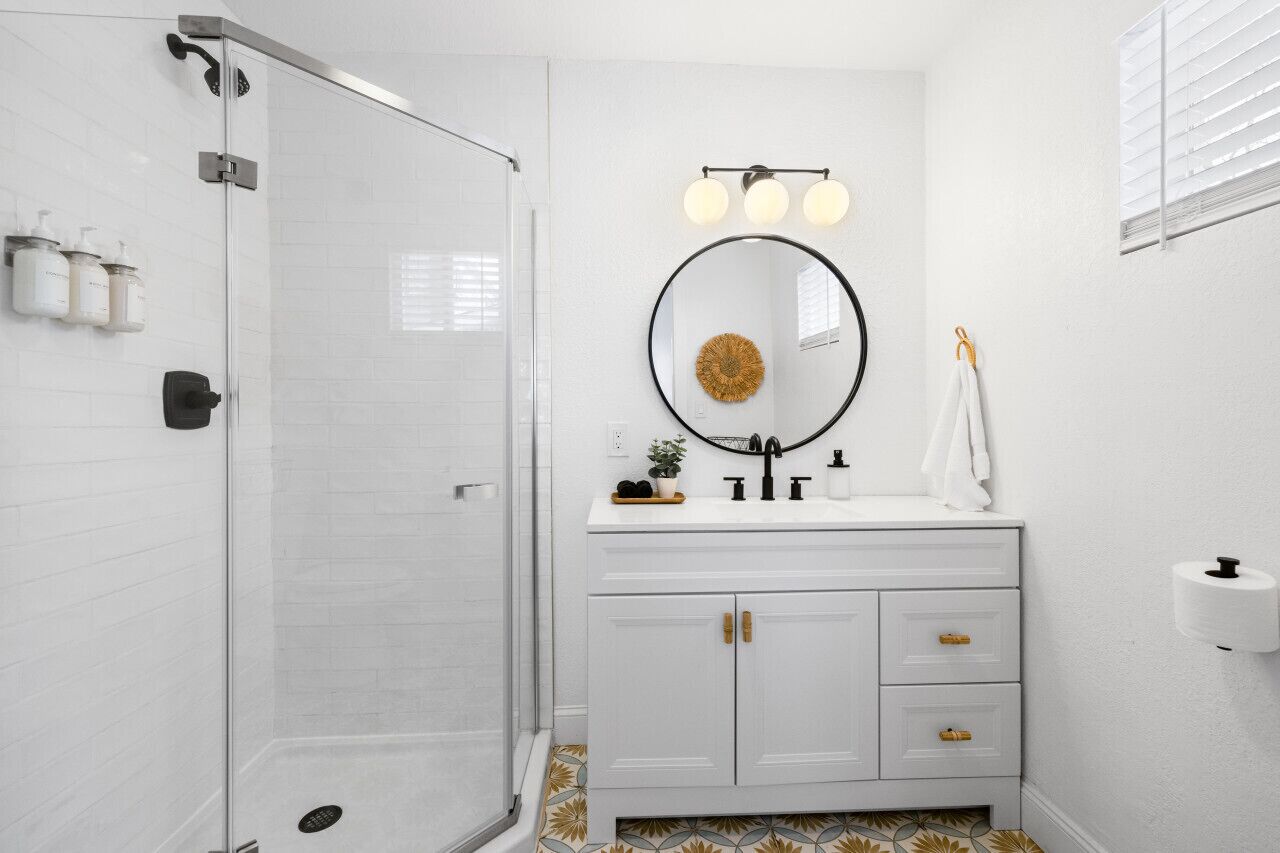 A bathroom with a glass shower enclosure, a white vanity with gold handles, a round mirror with a gold accent, and a towel rack.