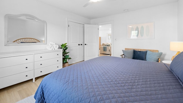 A bedroom with a bed, dresser, mirror, ceiling fan, and a door leading to another room.