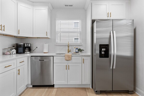 A modern kitchen with white cabinetry, a stainless steel refrigerator, a dishwasher, and a window with blinds.