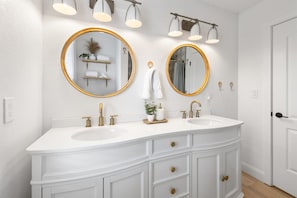 Ein helles Badezimmer mit zwei Waschbecken, goldenen Wasserhähnen und runden Spiegeln mit goldenen Rahmen.