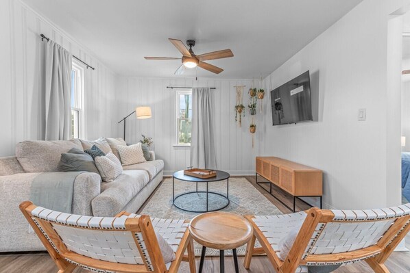Ein modernes Wohnzimmer mit einer Couch, zwei Sesseln, einem Couchtisch, einem Fernseher und einem Deckenventilator.