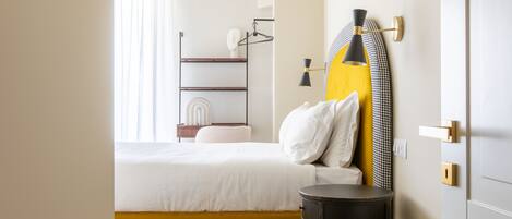 A hotel room with a bed, a bedside table, a lamp, a coat rack, and a window with curtains.