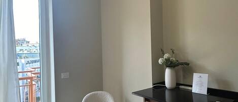 Habitación de hotel con suelo de madera en dibujo de espiga, una butaca tapizada de blanco, un mostrador de recepción con un jarrón de flores y vistas a edificios a través de un amplio ventanal.