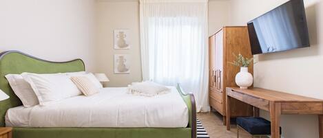 Un dormitorio con una cama verde, un escritorio de madera, un televisor en la pared y una ventana con cortinas.