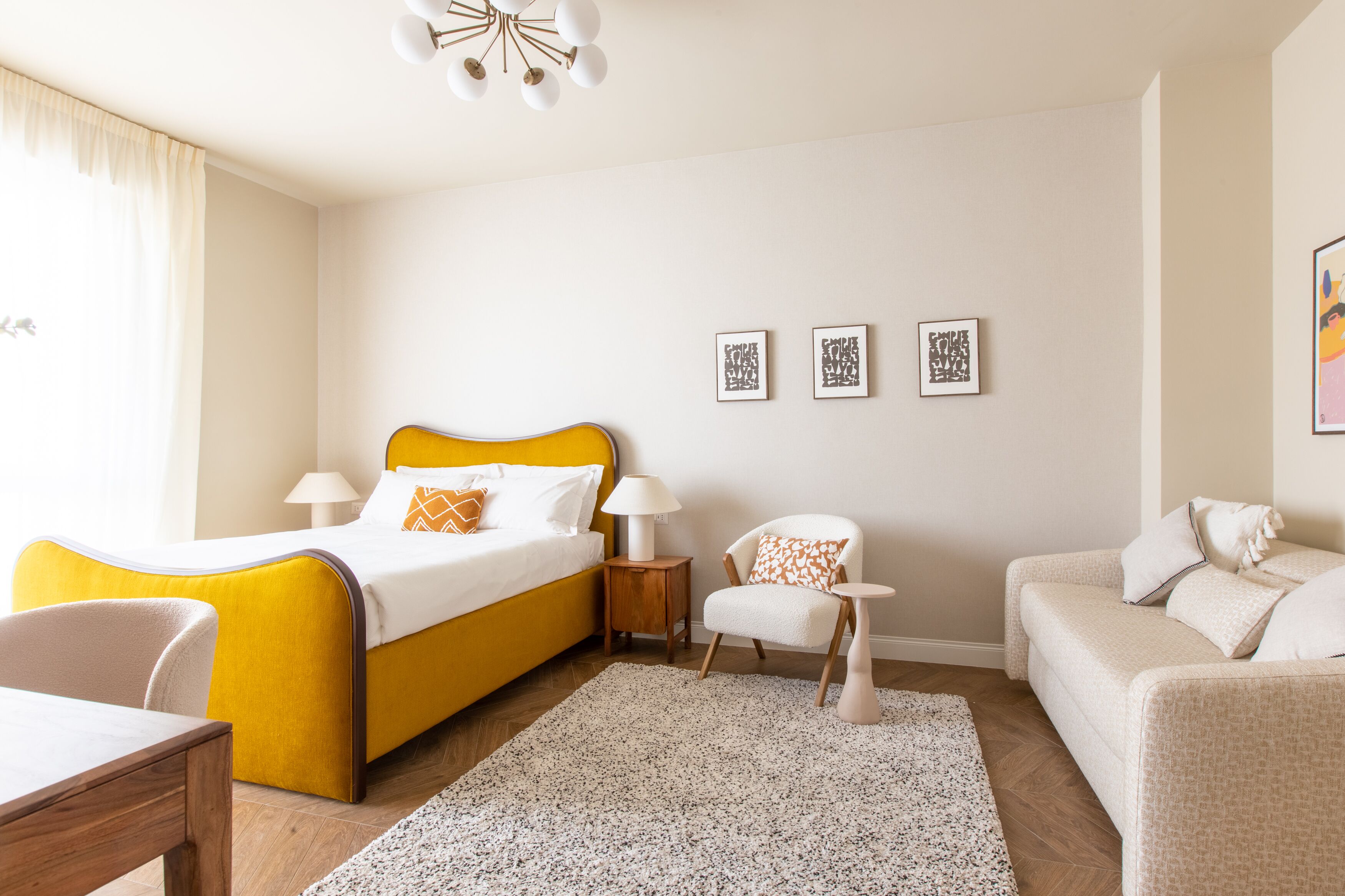 A hotel room with a yellow bed, a white armchair, a beige sofa, and a small round table. There are three framed pictures on the wall.