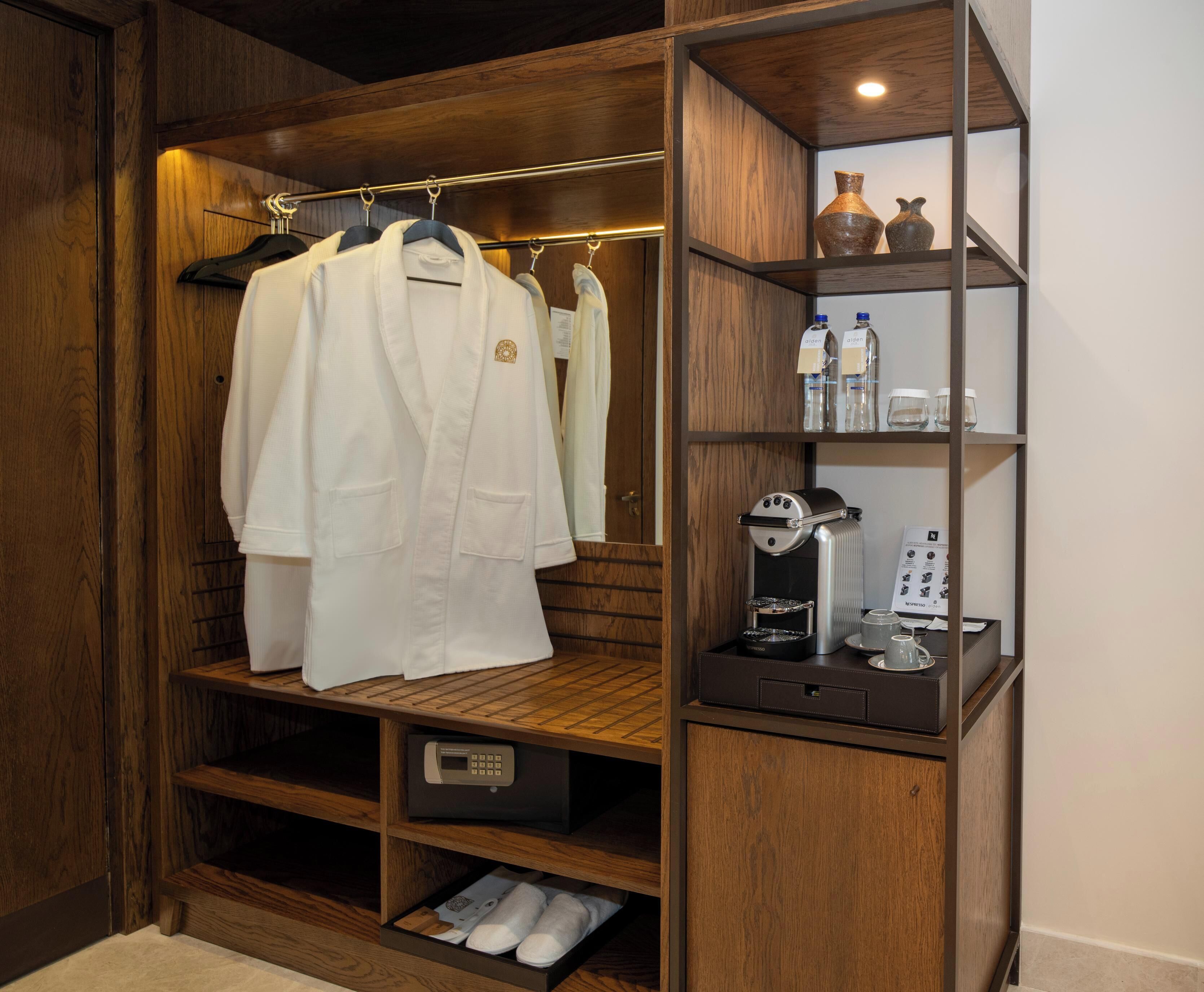 A hotel room with a wooden wardrobe, a hanging robe, a coffee machine, and a shelf with toiletries.