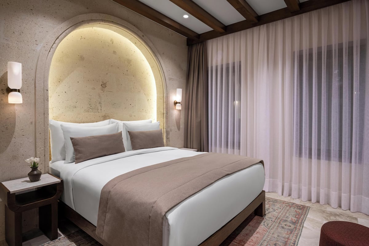 Habitación de hotel con una cama grande, una mesita de noche con un jarrón de flores y una ventana amplia con cortinas traslúcidas.