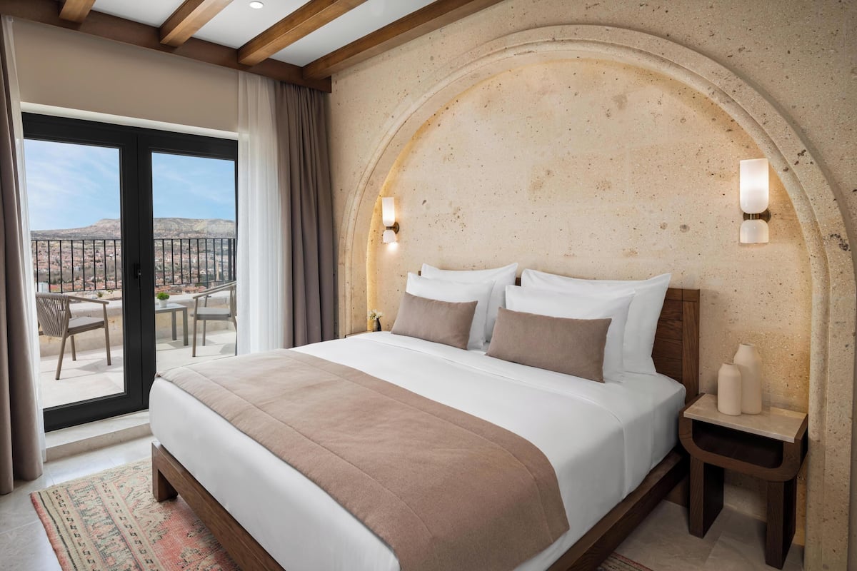 Habitación de hotel con una cama grande, una mesita pequeña, una mesita de noche y vistas al exterior.