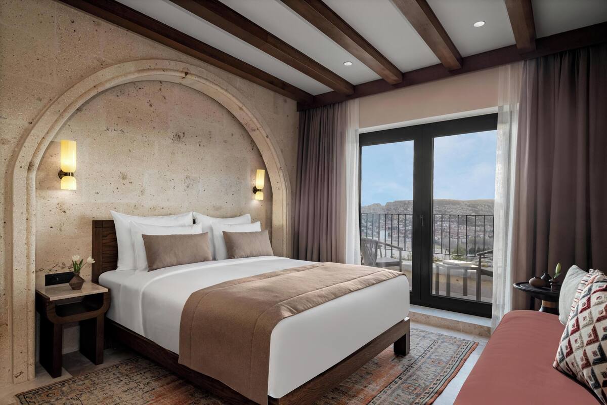 Un dormitorio amplio con una cama grande, una mesita de noche con un jarrón y vistas a la ciudad a través de un ventanal.