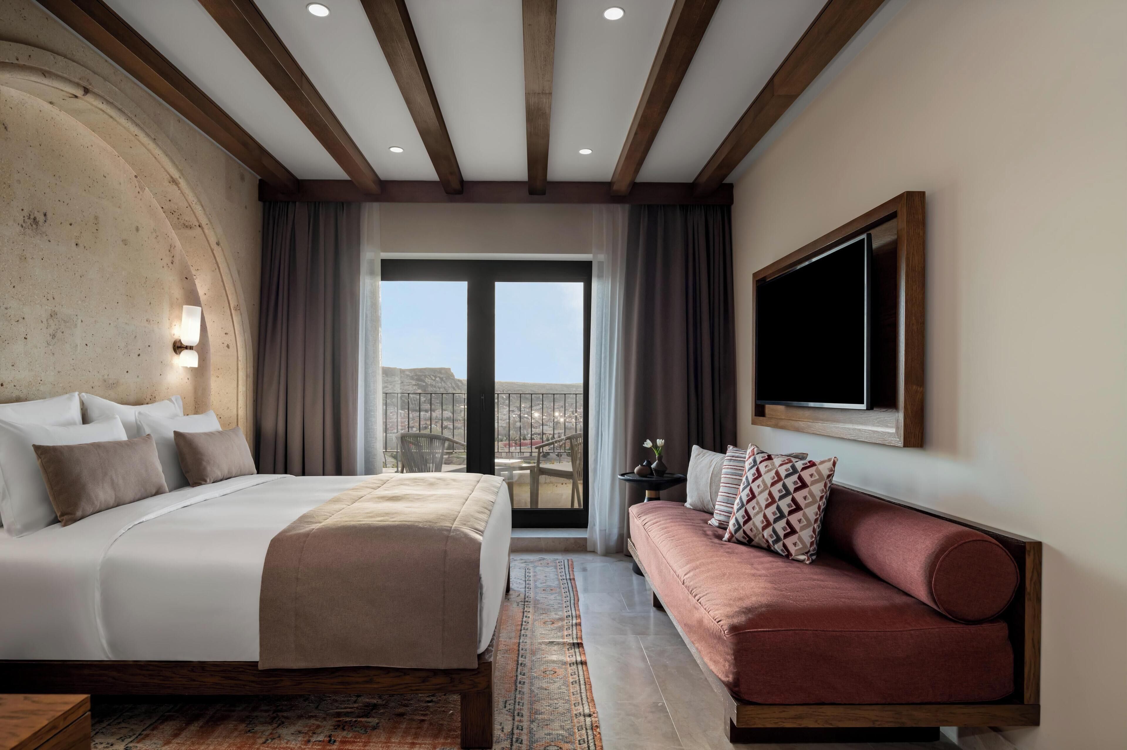 A hotel room with a large bed, a sofa, a TV, and a balcony with a view.