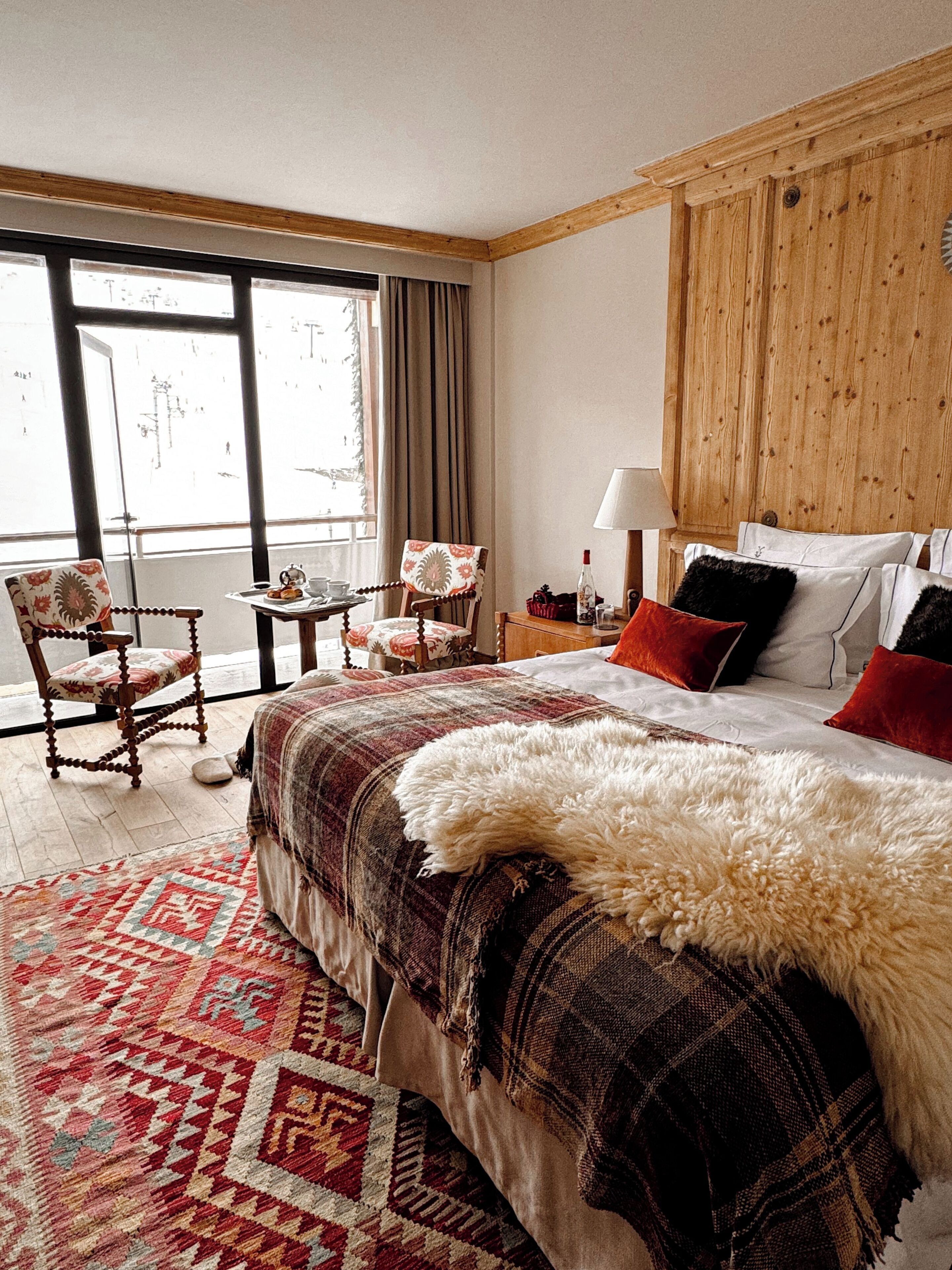 Habitación de hotel con una cama grande, un cabecero de madera, una ventana con cortinas, una mesita con lámpara y dos sillas.