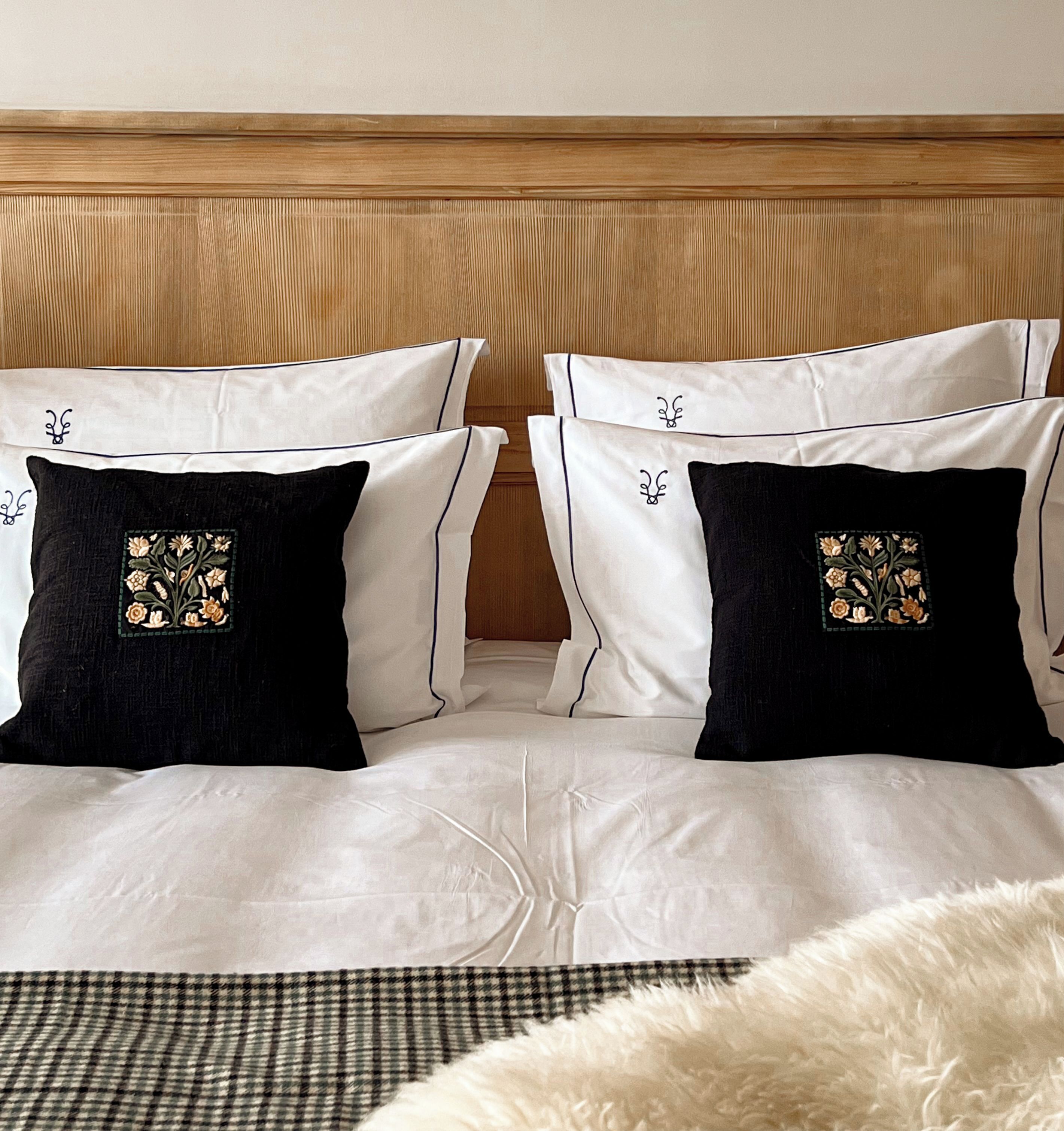 Una cama con ropa de cama blanca y dos almohadas negras con bordado floral, un cabecero de madera y una manta blanca mullida.