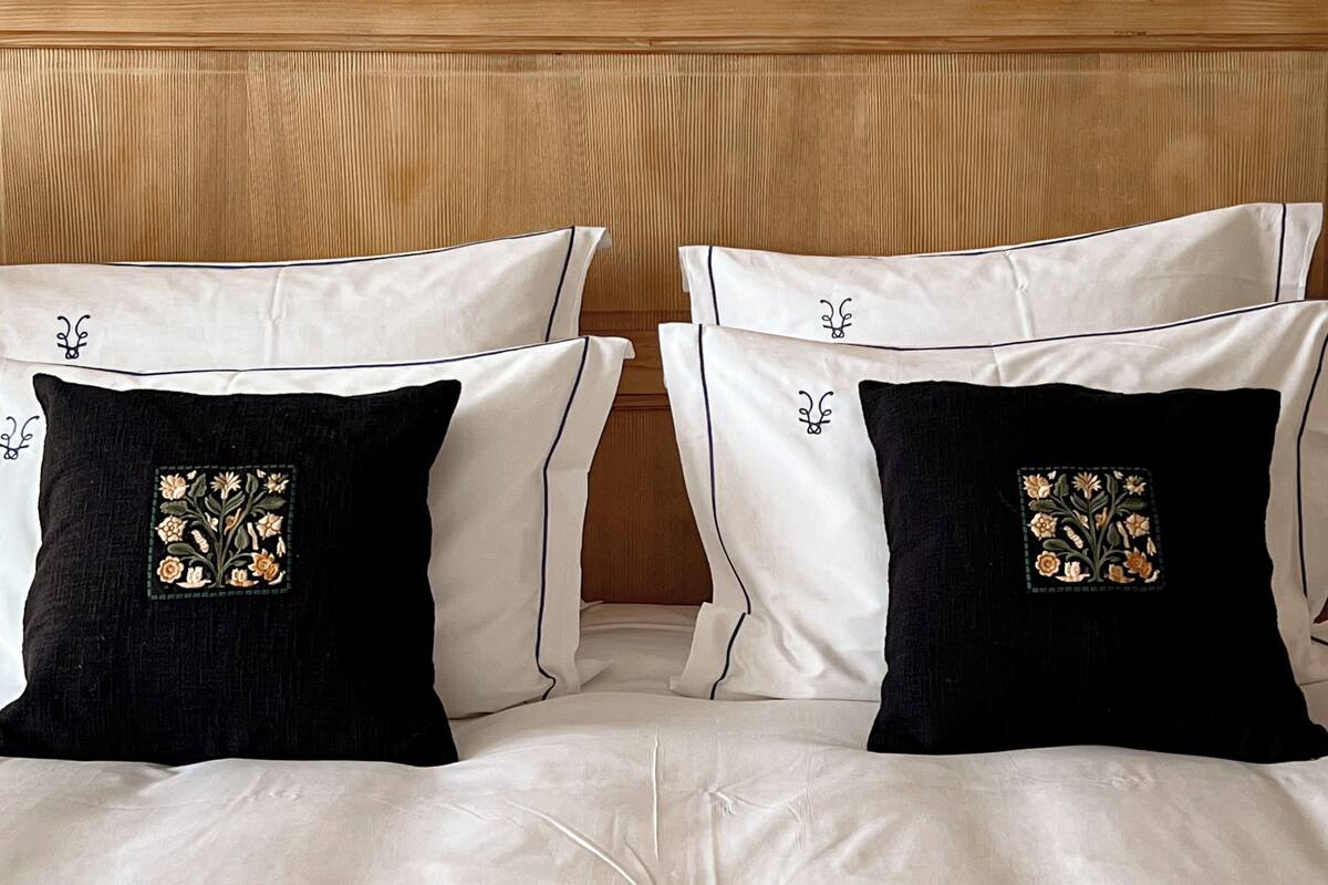 Ein Bett mit weißer Bettwäsche, zwei schwarze Kissen mit Blumenstickerei, ein hölzernes Kopfteil und eine flauschige, weiße Decke.