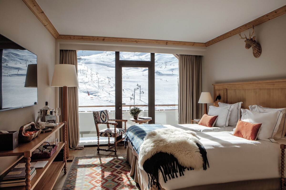 A hotel room with a large bed, a wooden headboard, a desk with a lamp, a chair, a TV, and a view of a snowy landscape.