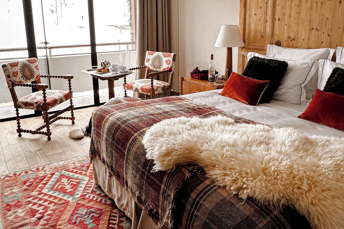 A hotel room with a large bed, a wooden headboard, a window with curtains, a small table with a lamp, and two chairs.
